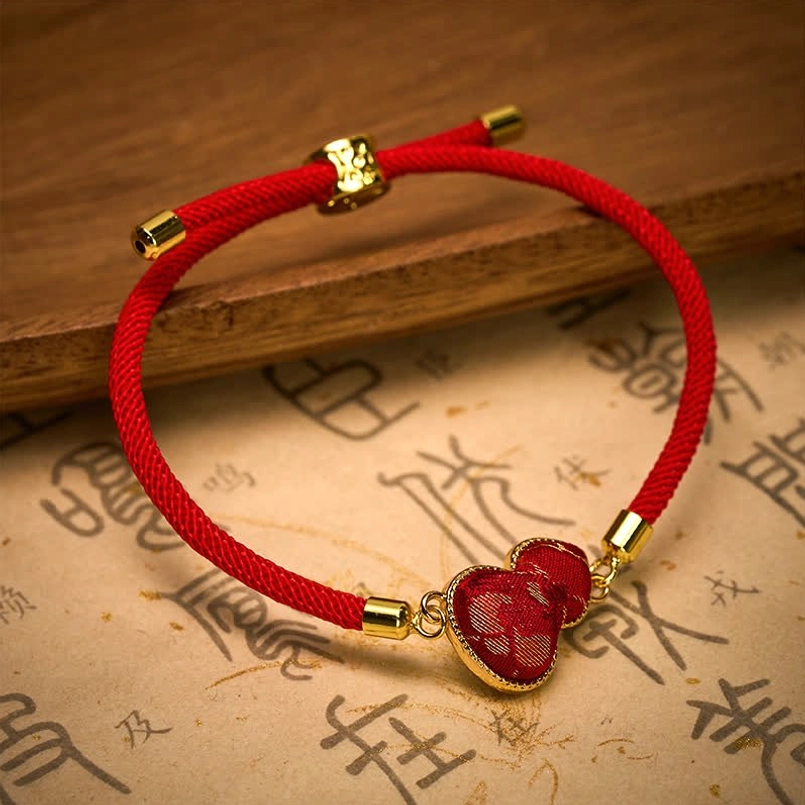 Red silk cord gourd charm bracelet with gold frame lying flat on wooden board