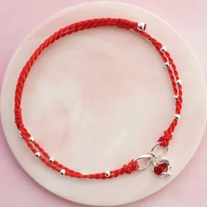 Red string infinity love anklet with heart pendant and silver beads on pink ceramic plate, flat lay view