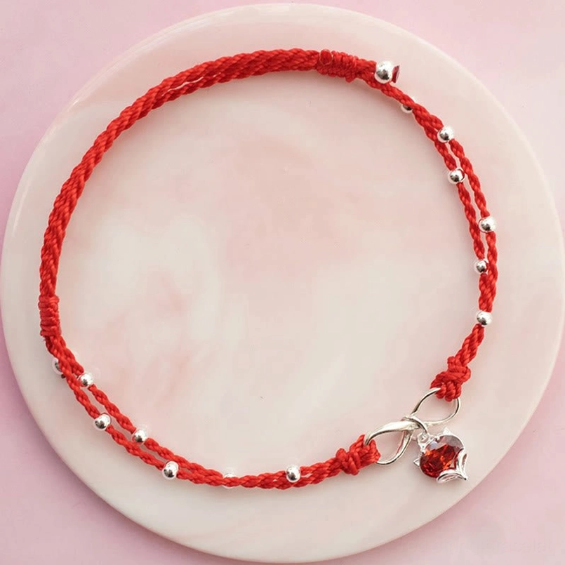 Red string infinity love anklet with heart pendant and silver beads on pink ceramic plate, flat lay view