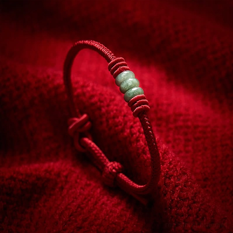 Handmade red string jade protection bracelet with braided cord and green jade beads on fabric background