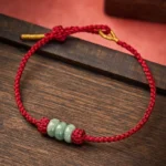 Red string jade bead bracelet with gold-tone cord ends displayed on wooden table with red background