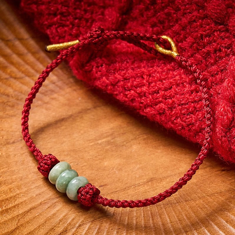 Handmade red braided jade bracelet resting on red knit fabric with gold metal fasteners