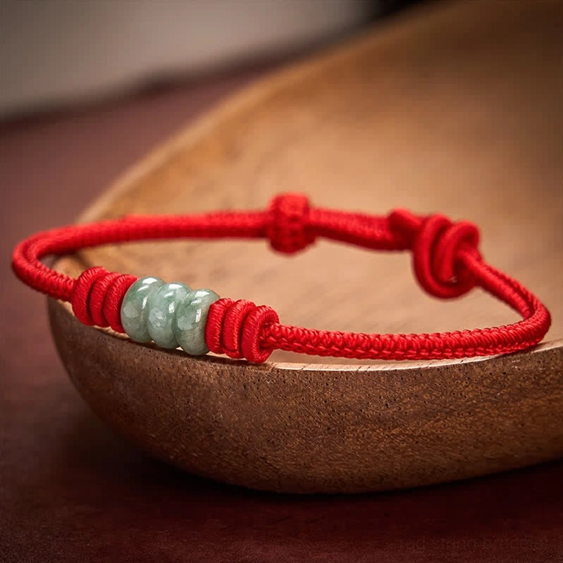 Adjustable red string jade bracelet with decorative knots displayed on wooden bowl background