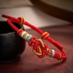 Red string jade heart knot bracelet with gold woven cord on dark wooden table