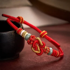Red string jade heart knot bracelet with gold woven cord on dark wooden table