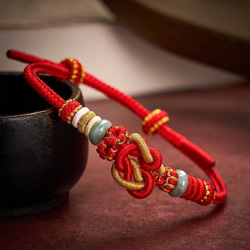 Red string jade heart knot bracelet with gold woven cord on dark wooden table