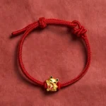 Red string lucky cat bracelet with golden charm on dark red fabric background - adjustable fortune bracelet