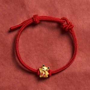 Red string lucky cat bracelet with golden charm on dark red fabric background - adjustable fortune bracelet