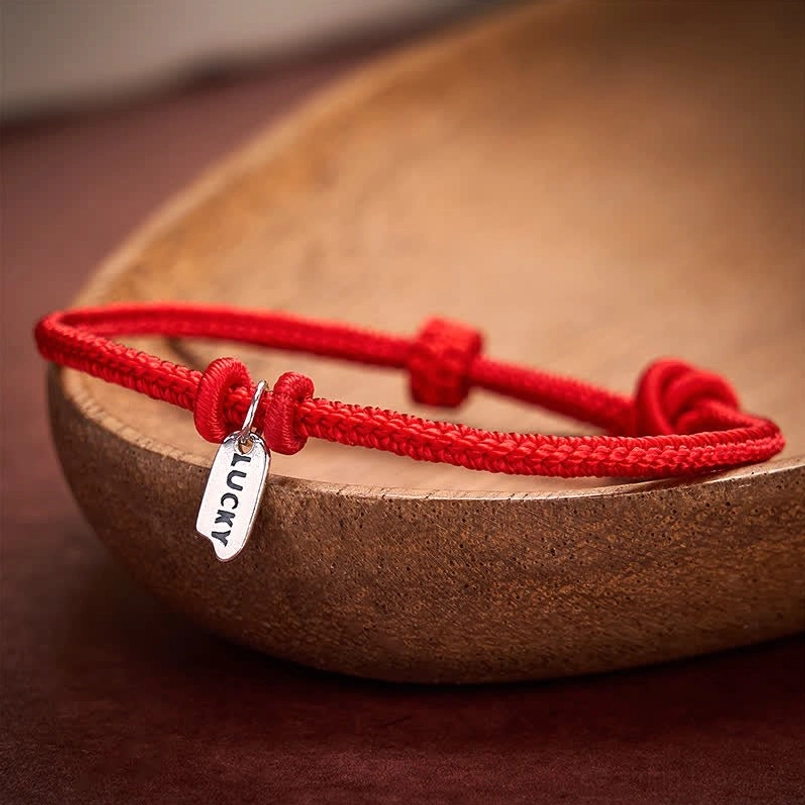 Red string bracelet with LUCKY metal tag and dual knots on wooden plate detail view