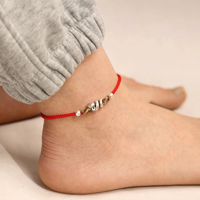 Red string snake anklet rear view on ankle featuring silver charms and decorative beaded accents against natural indoor background