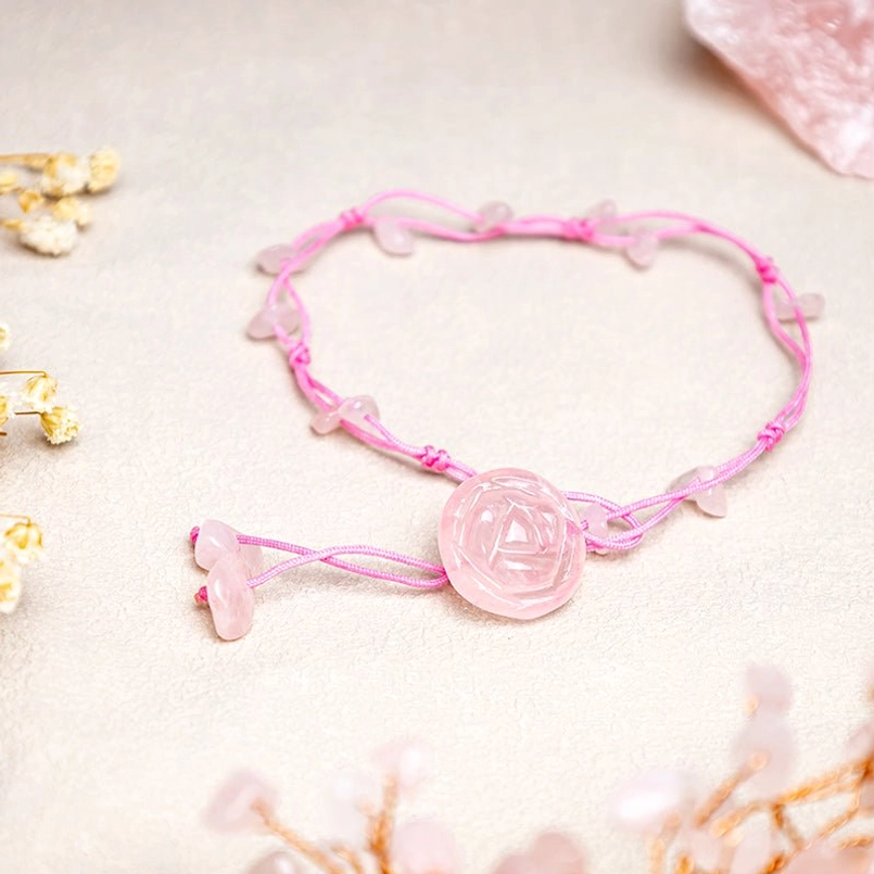 Rose quartz crystal anklet with pink braided cord and pink rose pendant on light table with dried flowers