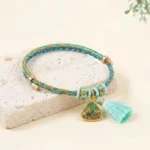Five-color Song Brocade red string bracelet with blue and yellow woven cord and turquoise tassel featuring lucky charm pendant on stone background