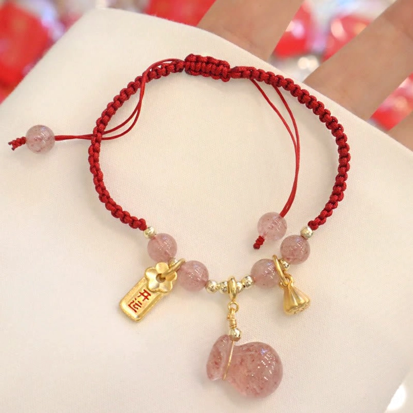 Handmade strawberry quartz braided red rope bracelet top down view with white fabric backdrop and red decorative accents showing lotus pod charm and lucky fortune tag