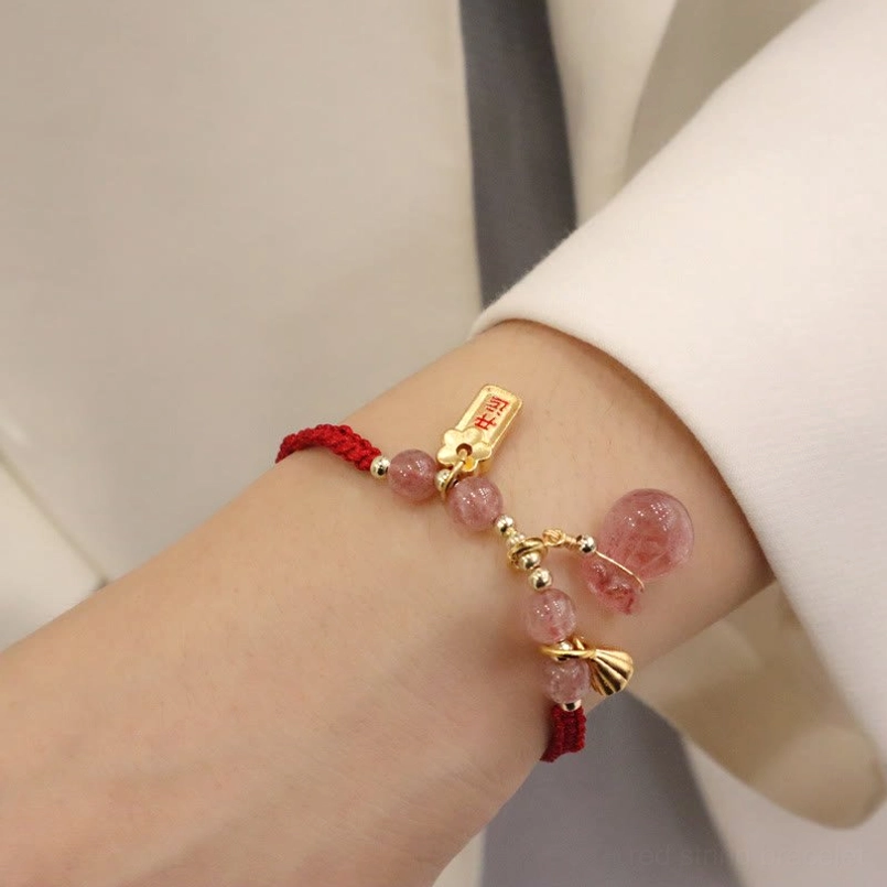 Worn strawberry quartz and red cord bracelet on wrist against light gray clothing showing lotus drop charm and fortune plaque pendant with braided rope details