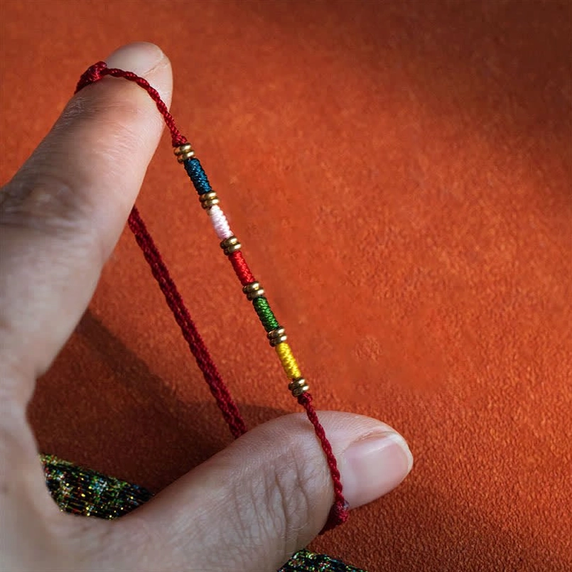 Close-up view of adjustable multicolor beaded red string bracelet held between fingers, showing colorful bead detail and cord craftsmanship