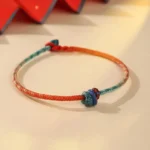 Hand-woven Tibetan red string bracelet with gradient teal color and decorative knots on beige background