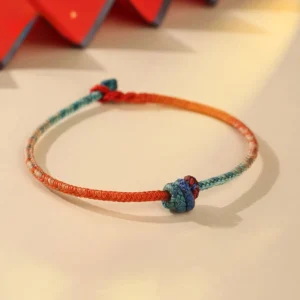 Hand-woven Tibetan red string bracelet with gradient teal color and decorative knots on beige background
