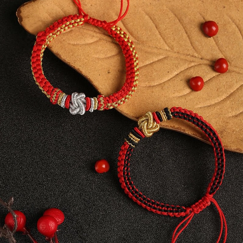 Two styles of red black and red gold string bracelets with adjustable cords displayed on wooden leaf