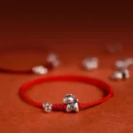 Year of the Horse sterling silver red string bracelet with bell charm close-up detail on reddish fabric background