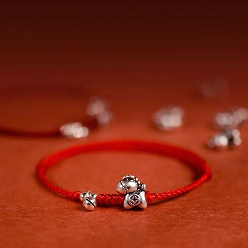 Year of the Horse sterling silver red string bracelet with bell charm close-up detail on reddish fabric background