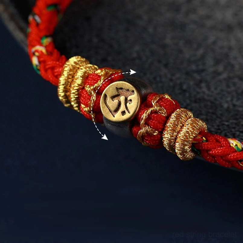 Macro close-up detail of zodiac natal buddha bracelet showing red woven cord, engraved gold mantra bead, and decorative gold knots